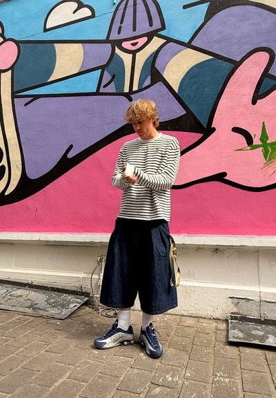 Camisa de manga larga a rayas en blanco y negro, pantalones cortos sueltos de color azul oscuro, calcetines blancos y zapatillas deportivas gris-azul, de pie frente a un mural vibrante.