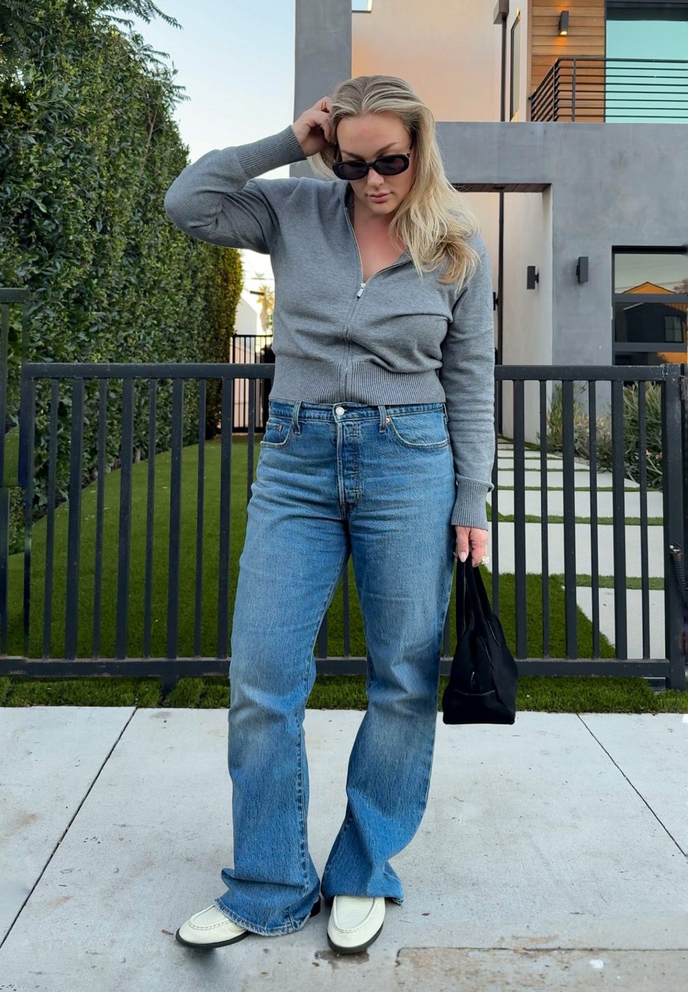 Femme portant des lunettes de soleil foncées, un pull gris zippé, un jean bleu à jambes larges et des chaussures blanches, tenant un sac à main noir, debout sur le trottoir.