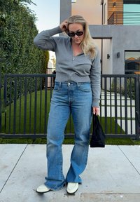 Femme portant des lunettes de soleil foncées, un pull gris zippé, un jean bleu à jambes larges et des chaussures blanches, tenant un sac à main noir, debout sur le trottoir.