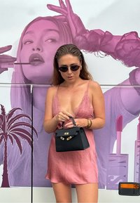 Svart håndveske med gullhardware, holdt av en person i en rosa satengkjole. Bakgrunnen har et stort rosa grafikk av en kvinne.