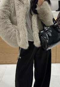 Umělá kožešinová bunda v světle béžové barvě s nadýchanou strukturou, kombinovaná s černými tepláky s bílými pruhy a černou koženou kabelkou.