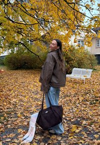 Brun skinnjakke, lyseblå jeans og en svart tote-veske med et pastellfarget skjerf. Høstblader dekker bakken i en park.