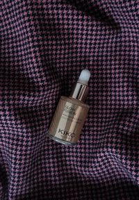 Flacon compte-gouttes en verre de KIKO Glow Fusion avec des gouttes illuminatrices, liquide ambre, capuchon argenté, posé sur un tissu à motif pied-de-poule en rose et bleu marine.