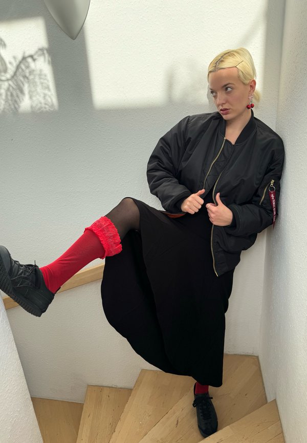 Čierna bomber bunda, čierna sukňa, červené návleky na nohy s čipkovým lemom a čierne tenisky. Zakrivené drevené schody v svetlom prostredí.