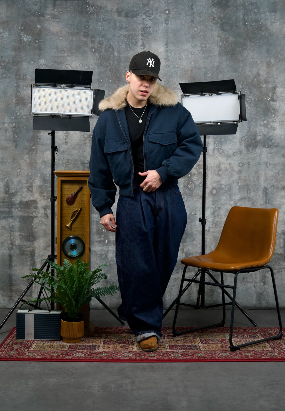 Jeune homme portant une veste bleu foncé et une casquette, debout sur un tapis rouge à motifs, entre deux éclairages de studio, une plante, une radio vintage et une chaise beige.