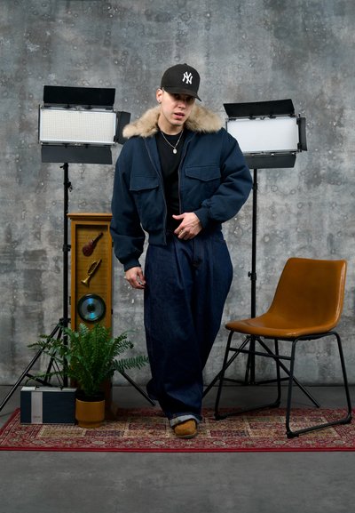 Joven con chaqueta azul oscuro y gorra de pie sobre una alfombra roja con diseño, entre dos luces de estudio, una planta, una radio vintage y una silla beige.