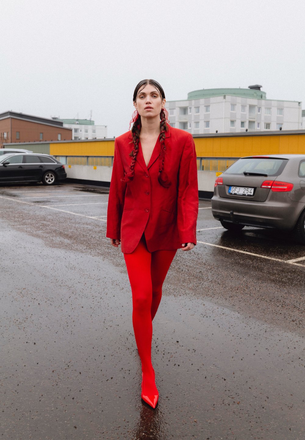 Rdeč blazer in ustrezne rdeče legice z ostrimi visokimi petami. Oblačilo ima elegantno zasnovo in je postavljeno proti sivi mestni ozadju.
