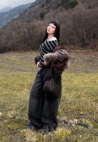 Joven de pie en un campo de hierba con una blusa de rayas sin hombros, chaqueta de piel, pantalones anchos negros y sosteniendo un bolso negro.