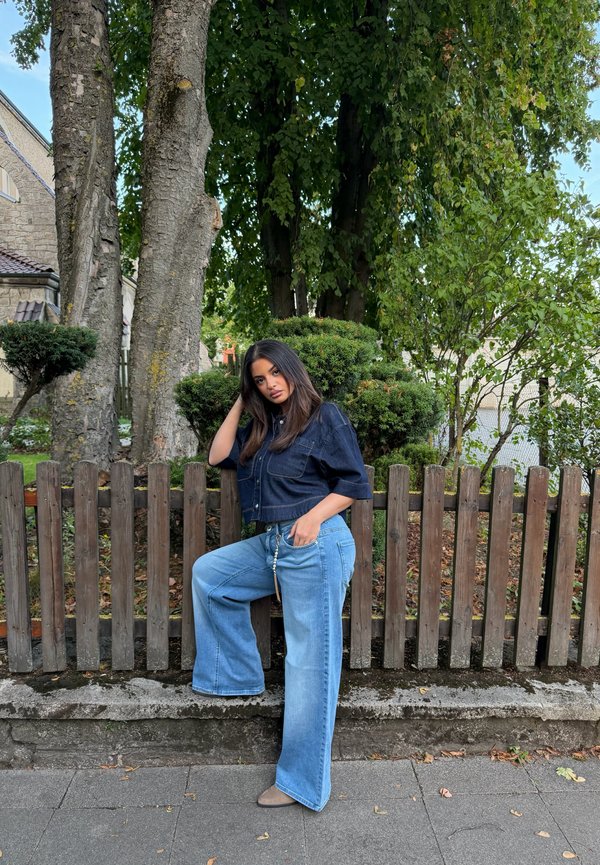 Jeune femme en haut foncé et jean large bleu posant avec un pied sur un mur bas en pierre devant une clôture en bois et des arbres.