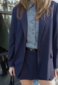 Tengerészkék csíkos blézer szürke gombos pulóver fölött, szűk szoknyával párosítva. Fekete öv és kézitáska teszi teljessé a megjelenést.