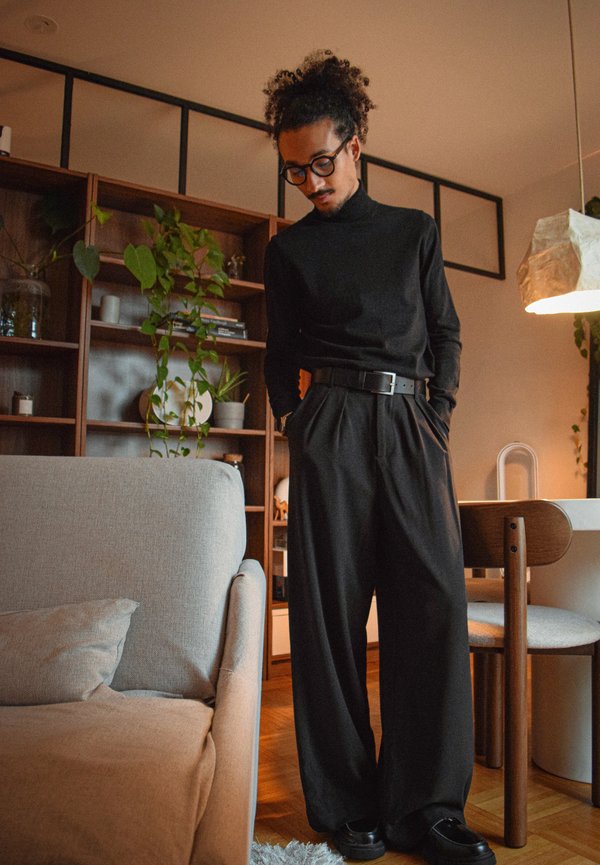Persona con cabello rizado, vestida con un cuello alto negro y pantalones anchos negros, de pie en una sala cerca de un sofá beige y estanterías de madera con plantas.