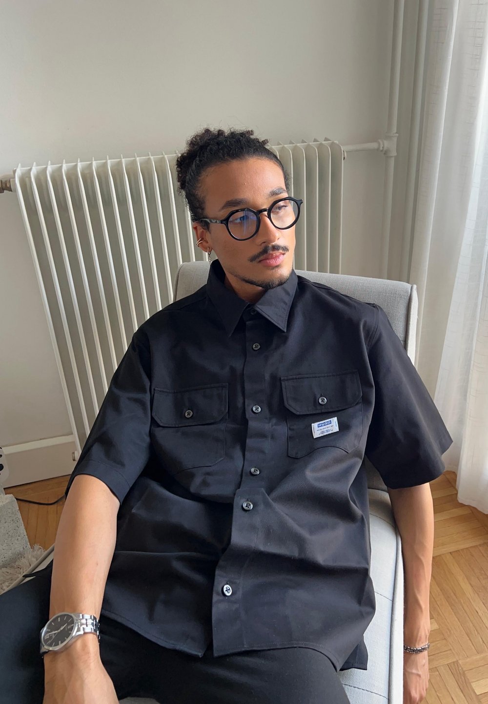 Zwart shirt met korte mouwen, voorzien van twee borstzakken en een label. Glad textuur van de stof. Model zit op een stoel met een neutrale achtergrond.
