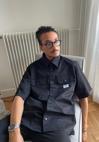Zwart shirt met korte mouwen, voorzien van twee borstzakken en een label. Glad textuur van de stof. Model zit op een stoel met een neutrale achtergrond.