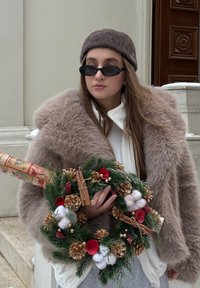 Manteau en fausse fourrure beige doux, haut blanc, tenant une couronne de fête avec des pommes de pin, des accents rouges et blancs, et un cadeau roulé. Lunettes de soleil foncées et chapeau.