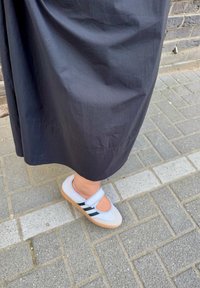 Persona que lleva una falda oscura y zapatos de tiras blancas con rayas negras, de pie sobre un pavimento de ladrillo gris cerca de una pared de ladrillo.
