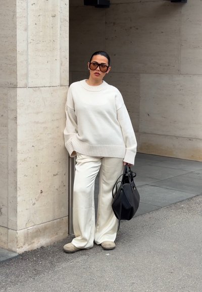 Mujer con suéter blanco de gran tamaño y pantalones, gafas de sol grandes, sosteniendo bolso negro, apoyada contra una pared de piedra clara al aire libre.