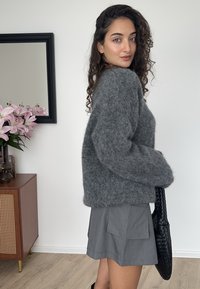 Pulover gri, pufos, cu formă dreptunghiulară, asortat cu o fustă scurtă plisată gri și o geantă tote neagră țesută. Texturi moi și design minimalist.