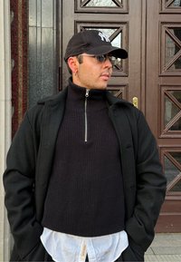 Homme portant une casquette noire, des lunettes de soleil, un sweat à capuche noir zippé, un manteau noir et une chemise blanche, debout devant une porte en bois aux motifs géométriques.