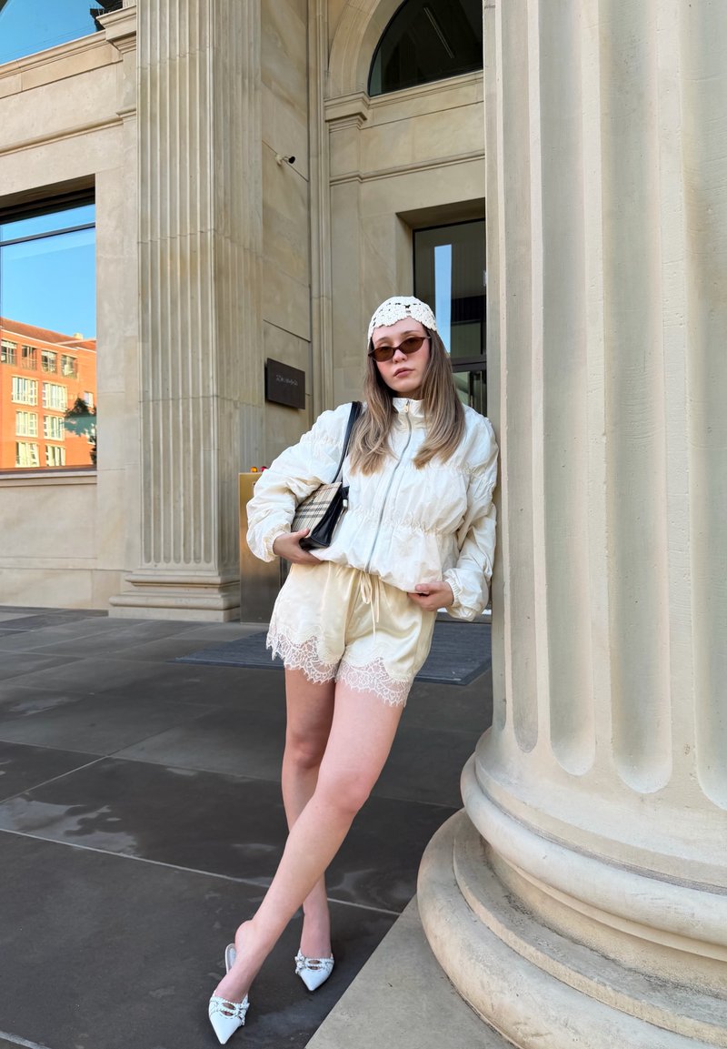 Jeune femme en shorts blancs bordés de dentelle et veste, foulard blanc sur la tête et lunettes de soleil, appuyée contre une grande colonne à l'extérieur d'un bâtiment.