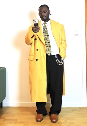 Hombre con abrigo largo amarillo, corbata estampada, pantalones anchos negros con cadenas, zapatos marrones, sosteniendo un teléfono inteligente y tomando una selfie en interiores.