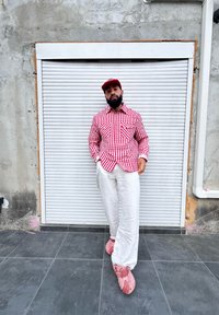 Camisa de botão vermelha e branca com riscas, calças de linho bege e tamancos cor-de-rosa. O fundo apresenta uma parede cinza e uma porta com persianas.