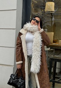 Ruda avies kailio striukė su balto kailio apykakle, baltas švelnios tekstūros šalikas, baltas viršus, šviesūs džinsai ir juoda rankinė. Dėvimos juodos akiniai nuo saulės.