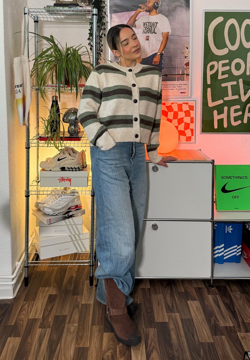 Knitted cream cardigan with green stripes and black buttons, light blue baggy jeans, brown ankle boots, indoor plants, and shelving.
