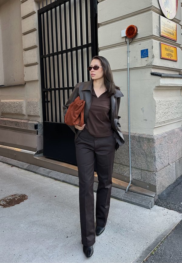 Giacca di pelle marrone sopra una camicia marrone e pantaloni abbinati. Porta una pochette color ruggine. Occhiali da sole scuri. Ambientazione urbana con un cancello.