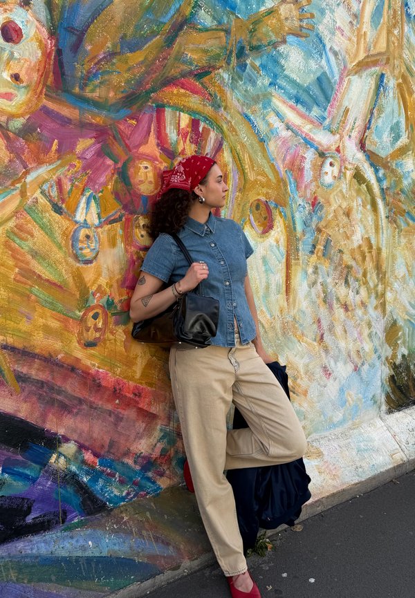 Jeune femme avec bandana rouge appuyée contre une fresque abstraite colorée, portant une chemise en denim, un pantalon beige et des chaussures rouges sur un trottoir urbain.