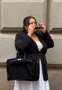 Sac à main en fourrure noire au design structuré avec des détails élégants en métal, associé à un blazer noir sur mesure porté sur une robe blanche.