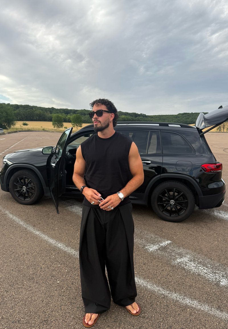 Homme portant des lunettes de soleil, un débardeur, un pantalon large et des sandales, debout près d'un SUV noir ouvert dans un grand parking extérieur.