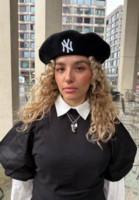 Svart ullberet med hvit NY-logo, kombinert med en svart skjorte med puffede ermer og hvit krage. Sølv anhengs halskjede synlig.