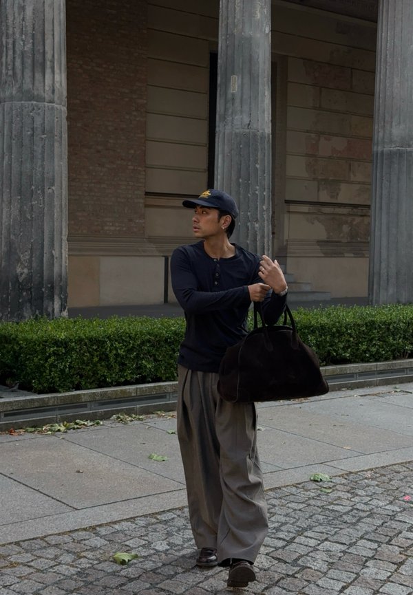 Mala de viagem em camurça preta, grande e macia, com formato arredondado. O modelo veste uma camisa preta de mangas longas e calças largas em cinza claro.