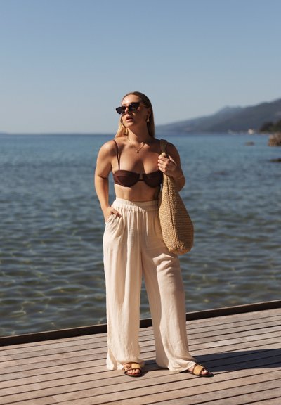 Top de bikini marrón, pantalones de pierna ancha color crema y un bolso de hombro tejido beige. De pie junto al agua, usando gafas de sol y sandalias planas.