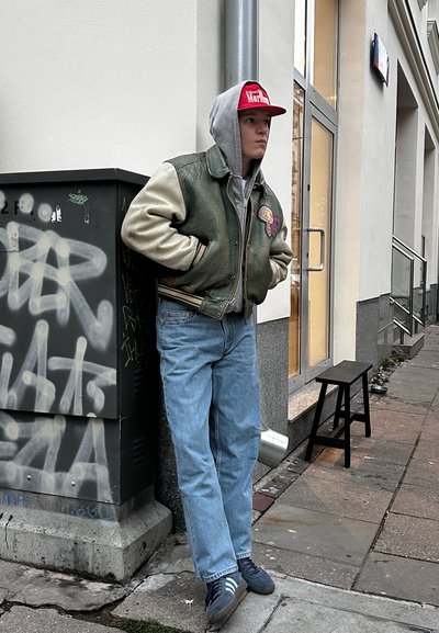 Chaqueta bomber de cuero verde con mangas crema, sudadera gris, jeans azules, zapatillas azules, gorra roja, de pie junto a una caja de servicios cubierta de graffiti.
