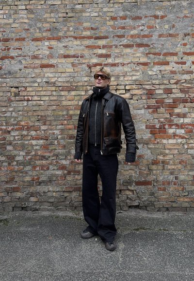 Hombre con chaqueta de cuero negra, suéter oscuro de punto, pantalones anchos negros y gafas de sol, de pie contra una pared de ladrillos sobre suelo de asfalto.