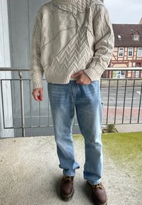 Pull beige avec des motifs texturés, y compris des tresses et des carreaux ; associé à un jean bleu clair et des chaussures marron. Se tenant sur un balcon.