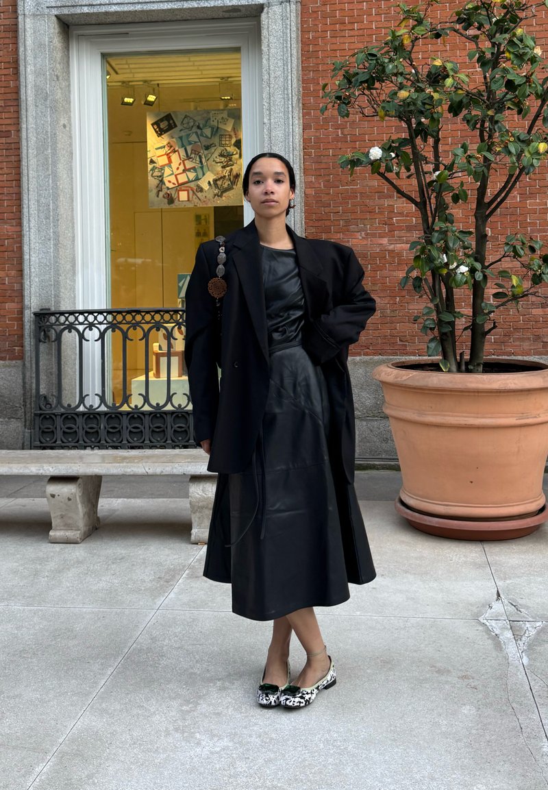 Femme portant une robe en cuir noire et un blazer noir oversize se tient à l'extérieur près d'une grande plante en pot et d'un banc devant un bâtiment en briques.