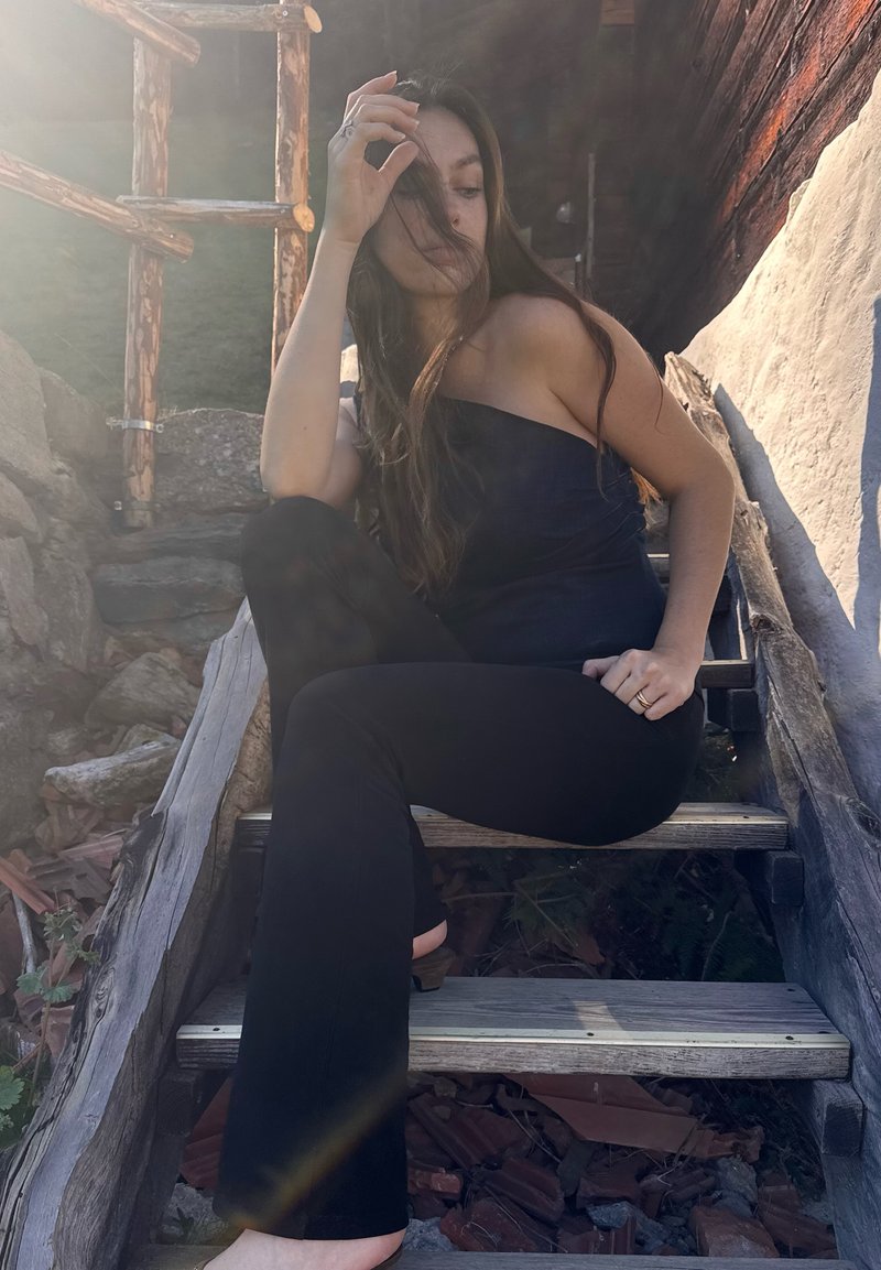 Zwarte slim-fit broek en een strapless donker top worden gedragen terwijl er op houten trappen wordt gezeten. Het zonlicht creëert een zachte gloed om de figuur.