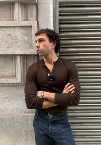 Moški s utrjenimi lasmi v rjavem dolgih rokavih in kavbojkah stoji s prekriženimi rokami, gleda levo, ob betonski steni in kovinski shutter ozadju.