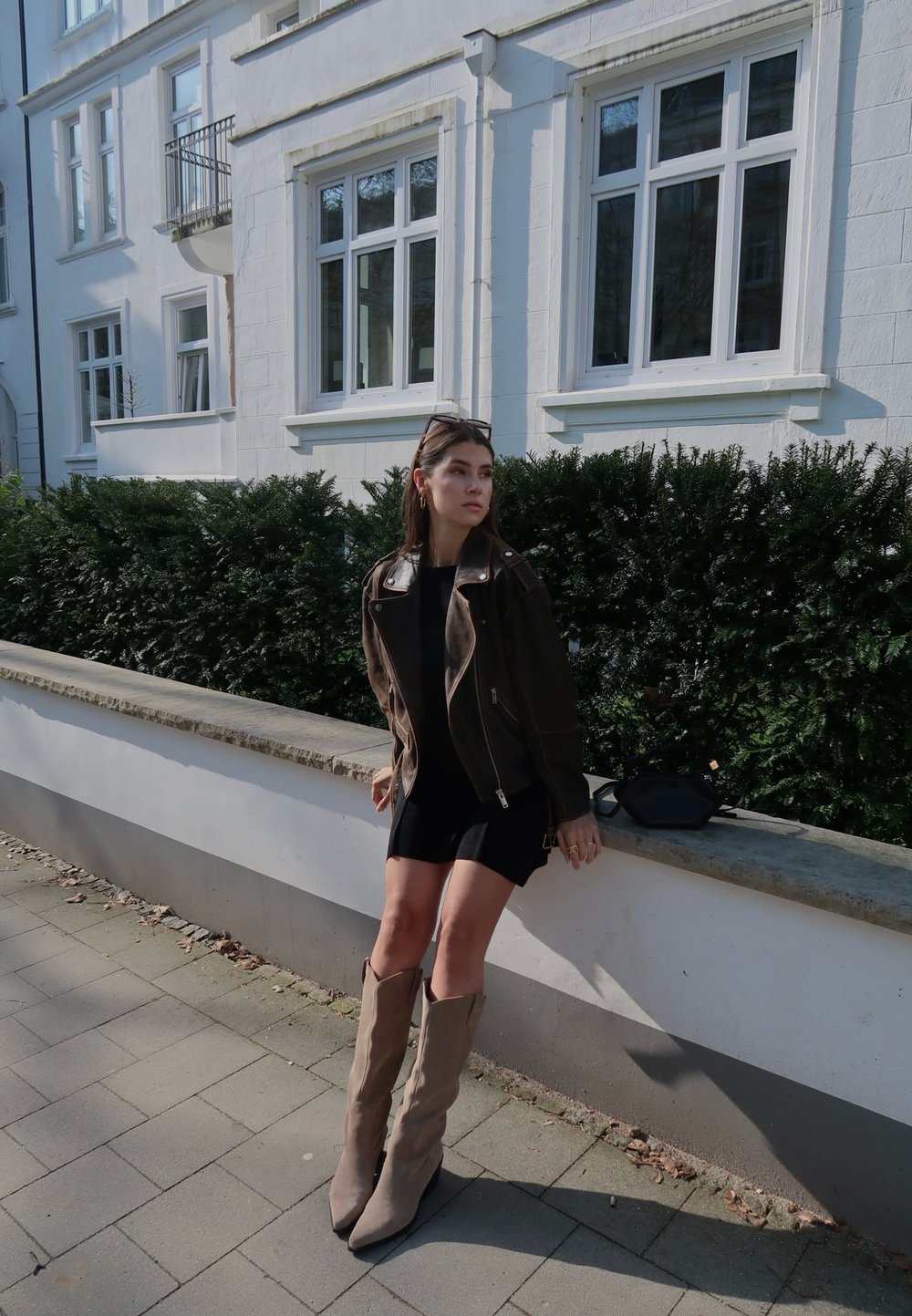 Rjava usnjena motoristična jakna, črna obleka, bež škornji do kolen s koničastimi prsti, stoji na kamnitem zidu blizu zelenih grmov.