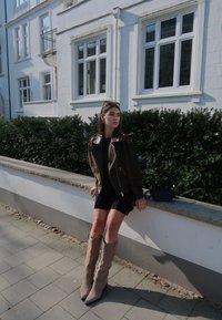 Rjava usnjena motoristična jakna, črna obleka, bež škornji do kolen s koničastimi prsti, stoji na kamnitem zidu blizu zelenih grmov.