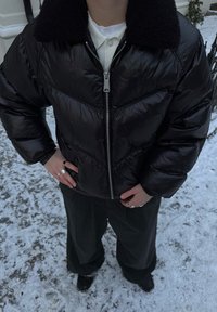 Veste matelassée noire avec col en fourrure noire texturée, dotée d'une fermeture éclair à l'avant et de détails de coutures géométriques, portée sur un pantalon foncé.