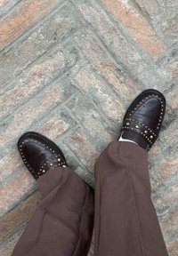 Mocassins en cuir marron avec des clous en laiton, portés avec un pantalon marron, debout sur un pavement en briques texturé et à motifs.