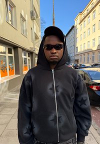 O persoană tânără îmbrăcată cu un hanorac negru cu motive în formă de stea, cu o șapc�ă de baseball și ochelari de soare, stând pe trotuarul unei zone urbane însorite, cu mașini parcate în apropiere.