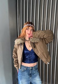 Personne portant une veste à col en fourrure marron, un haut bleu à paillettes, un jean, un diadème argenté et des colliers superposés, posant les yeux fermés et la main près du visage.