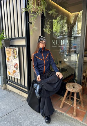Veste marine avec des rayures orange, pantalon noir surdimensionné, bottes noires et un bonnet noir avec du texte blanc. Le cadre comprend l'entrée d'un café et des plantes.