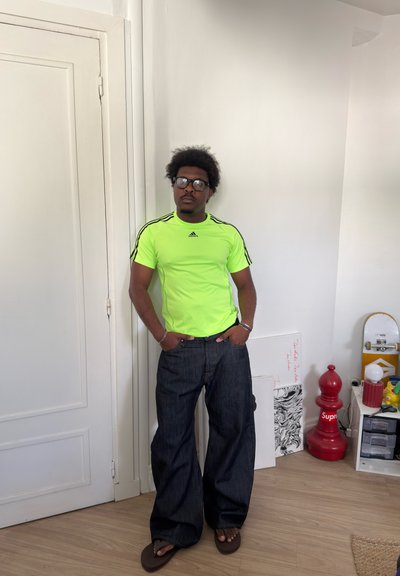 Hombre con camiseta Adidas verde neón y vaqueros oscuros y anchos, de pie con las manos en los bolsillos en una habitación con una pieza de ajedrez roja y una patineta cerca.