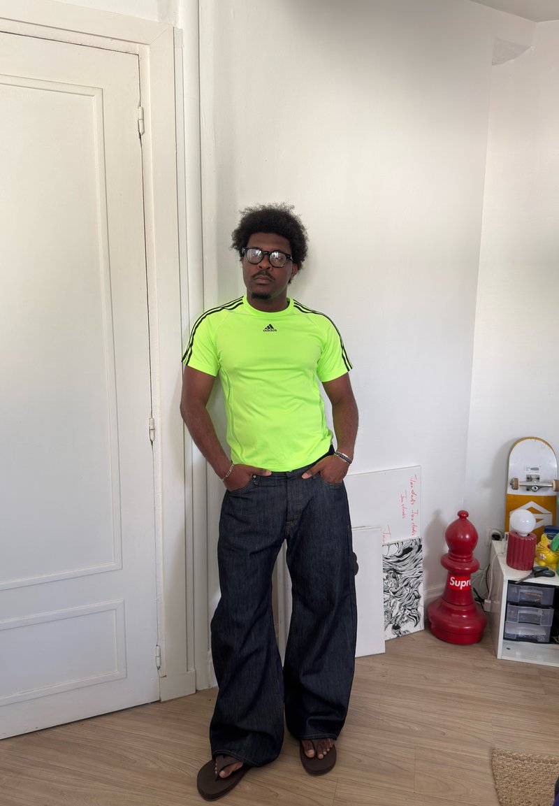 Mann iført neongrønn Adidas-skjorte og vide mørke jeans står med hendene i lommene i et rom med en rød sjakkbrikke og skateboard i nærheten.
