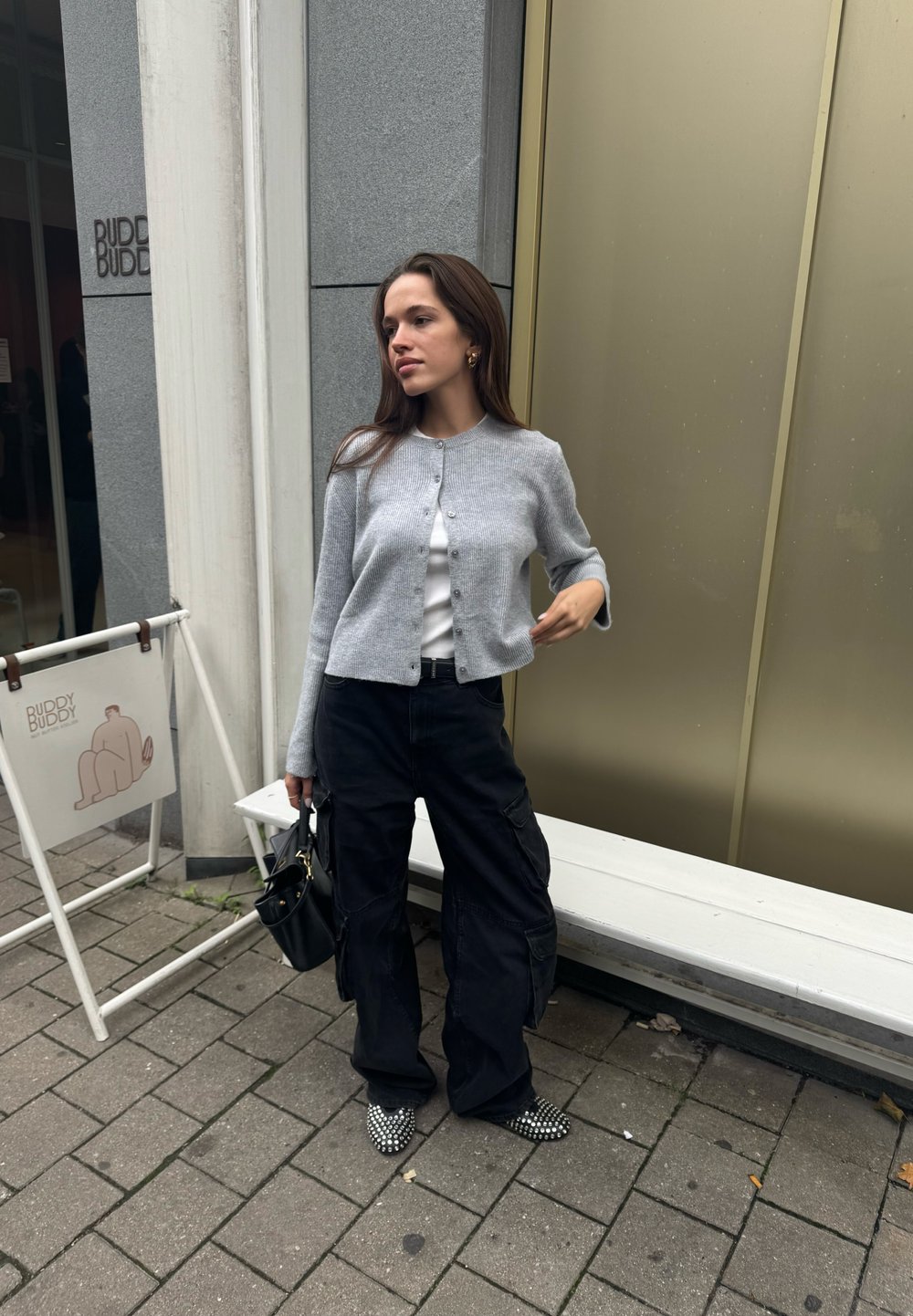 Cardigan acentuado cinza sobre uma blusa branca, combinado com calças de carga pretas. Sapatos de bico fino em preto e branco, levando uma pequena bolsa preta.
